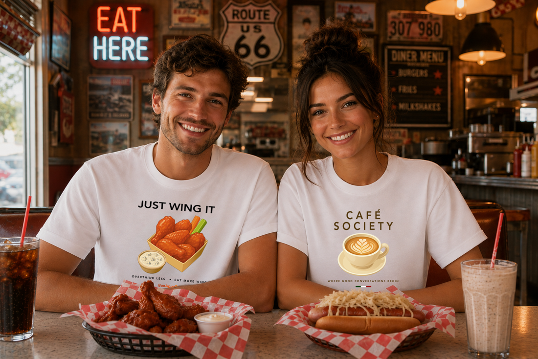 Couple wearing BonAppaTee graphic T-shirts enjoying chicken wings and a New York-style hot dog at a classic Route 66 American diner.